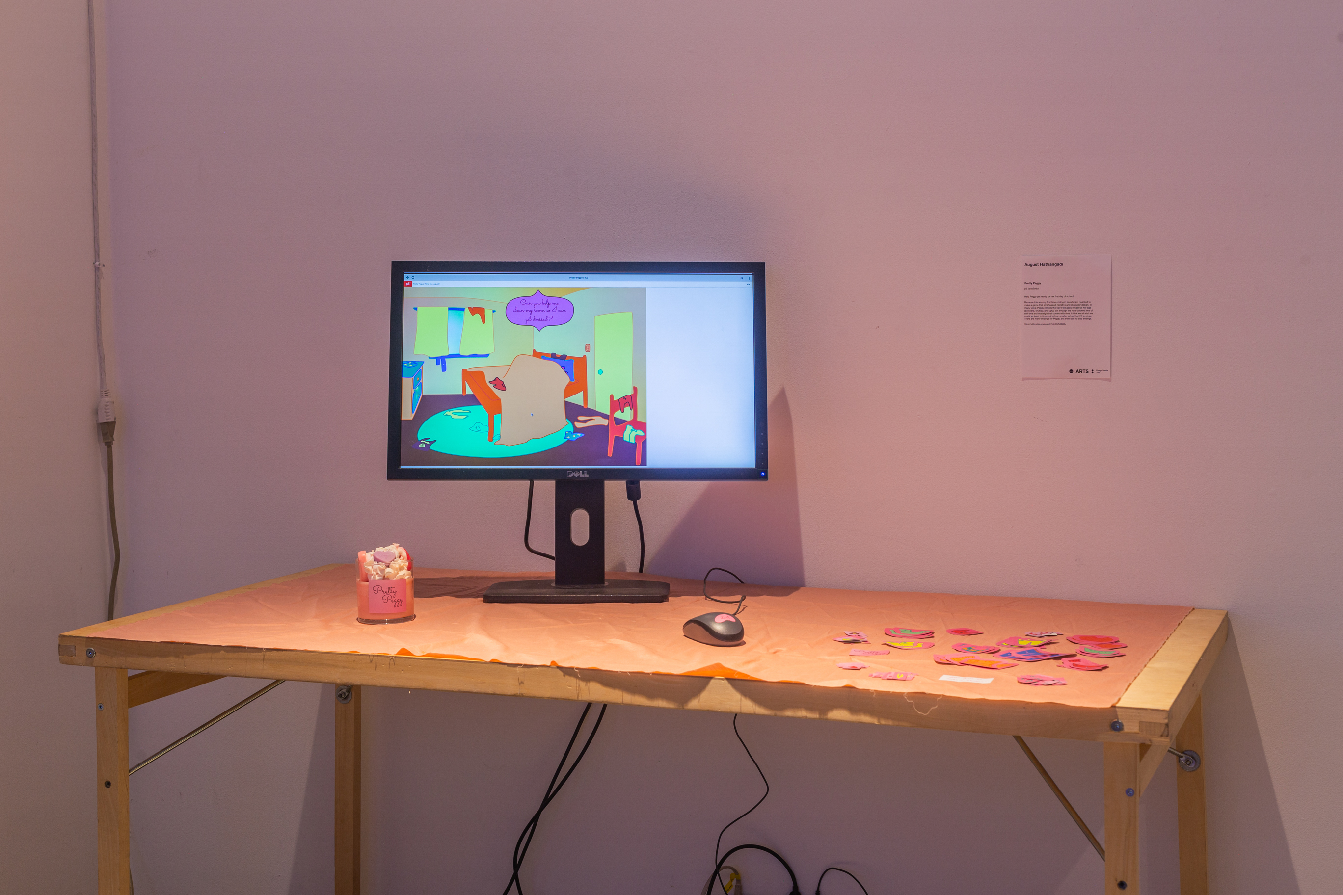 A table in a white space with a computer, a mouse, and a candle with candy in it. The computer has an interactive art piece displayed.